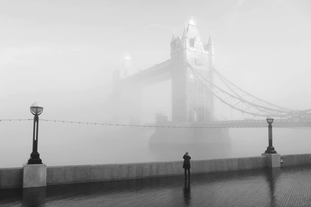 A person stands on the wet riverside walkway, gazing at London’s Tower Bridge, which is partially obscured by thick morning fog.